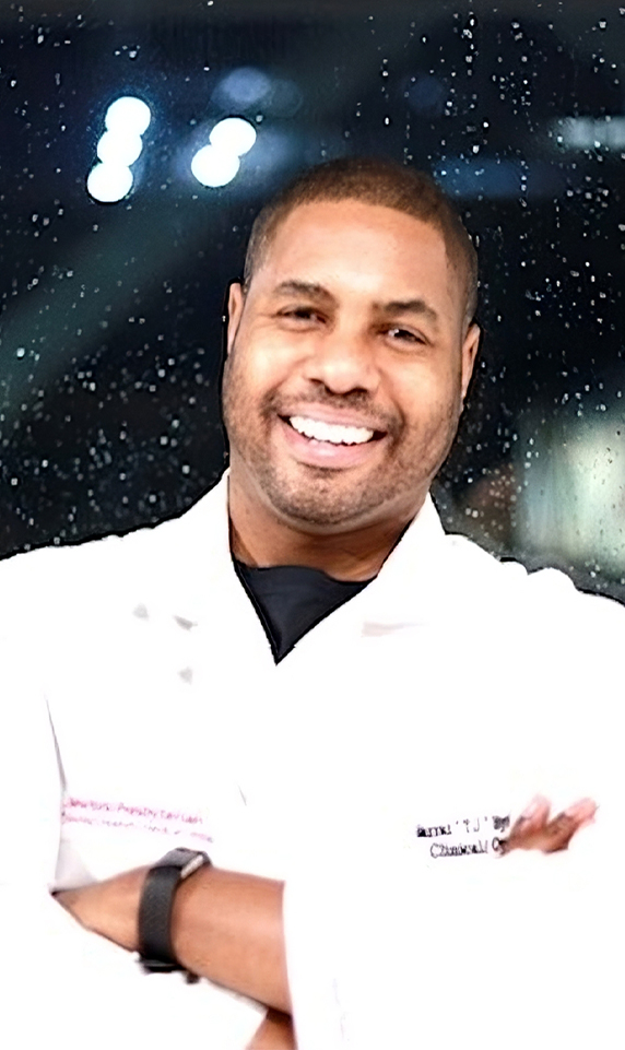 A headshot-style image of pharmacist Charrai Byrd. He is a Black man and is wearing a white lab coat while standing with his arms crossed in front of a window at night.