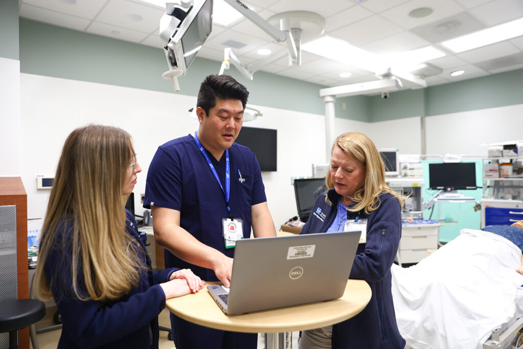 A candid image of pharmacist Alex Kim working on his laptop while being assisted by two other healthcare professionals. They are standing and looking at the laptop while in a room with a patient.