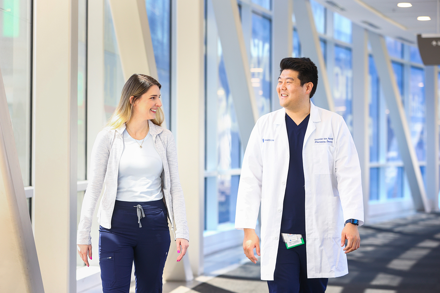 A candid image of pharmacist Alex Kim walking through a hallway in a healthcare setting with a patient. He is wearing a white lab coat over his blue scrubs.