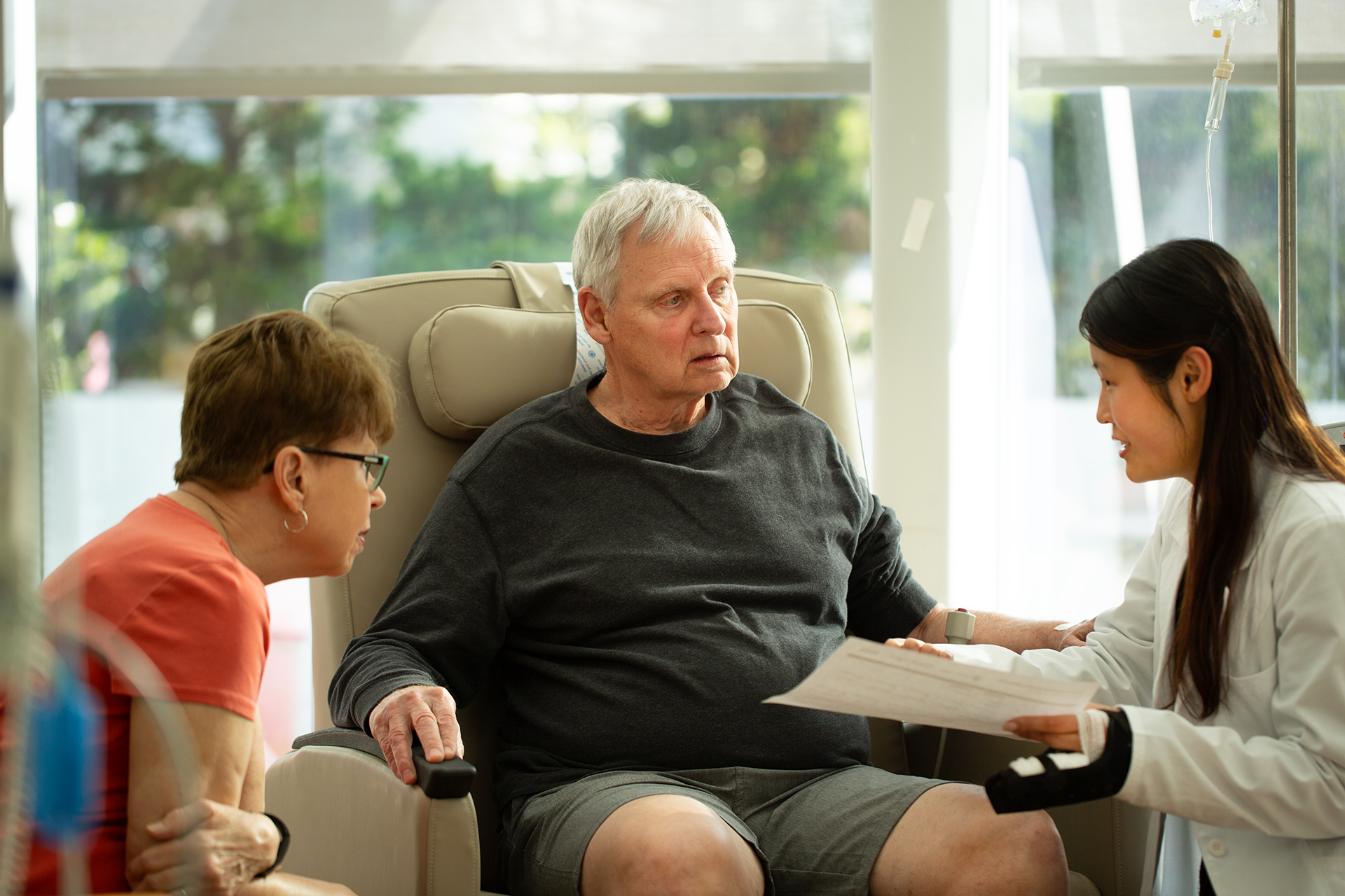 A health-system pharmacist showing a document to a patient and the patient's partner. The pharmacist is an Asian woman with long, dark hair, and the patient is an older caucasian man in a sweatshirt.