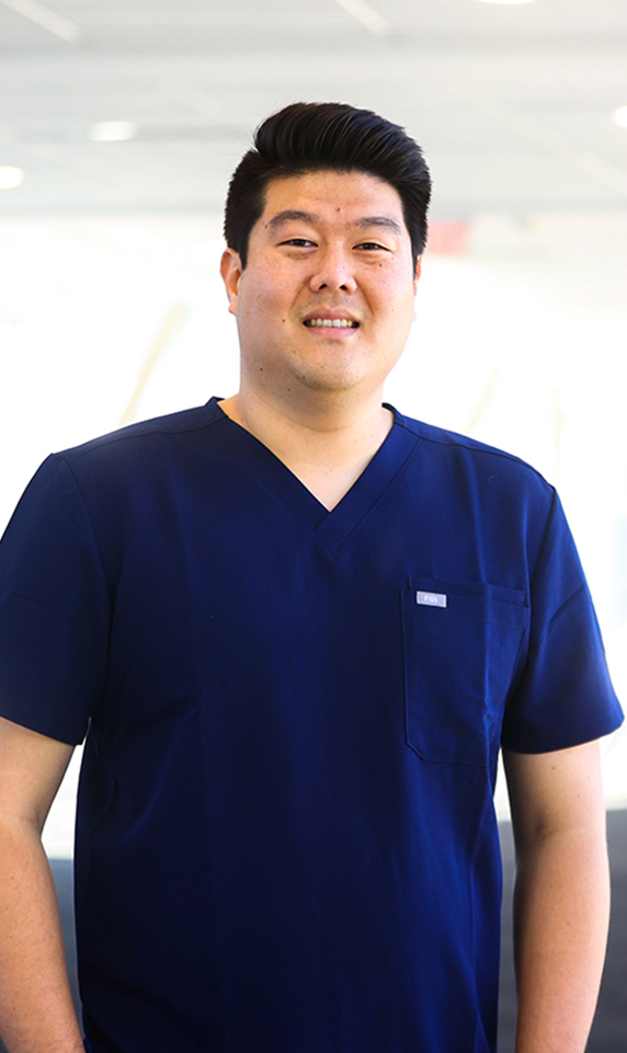 A headshot-style image of pharmacist Alex Kim. He is an Asian man with short, dark hair and is wearing dark blue scrubs.