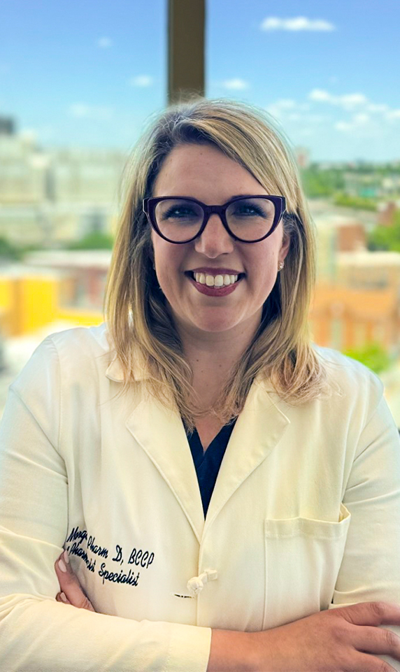 A headshot-style image of pharmacist Olivia Morgan. She has fair skin and medium-length, blonde hair, and is wearing thick-rimmed glasses and a white lab coat.