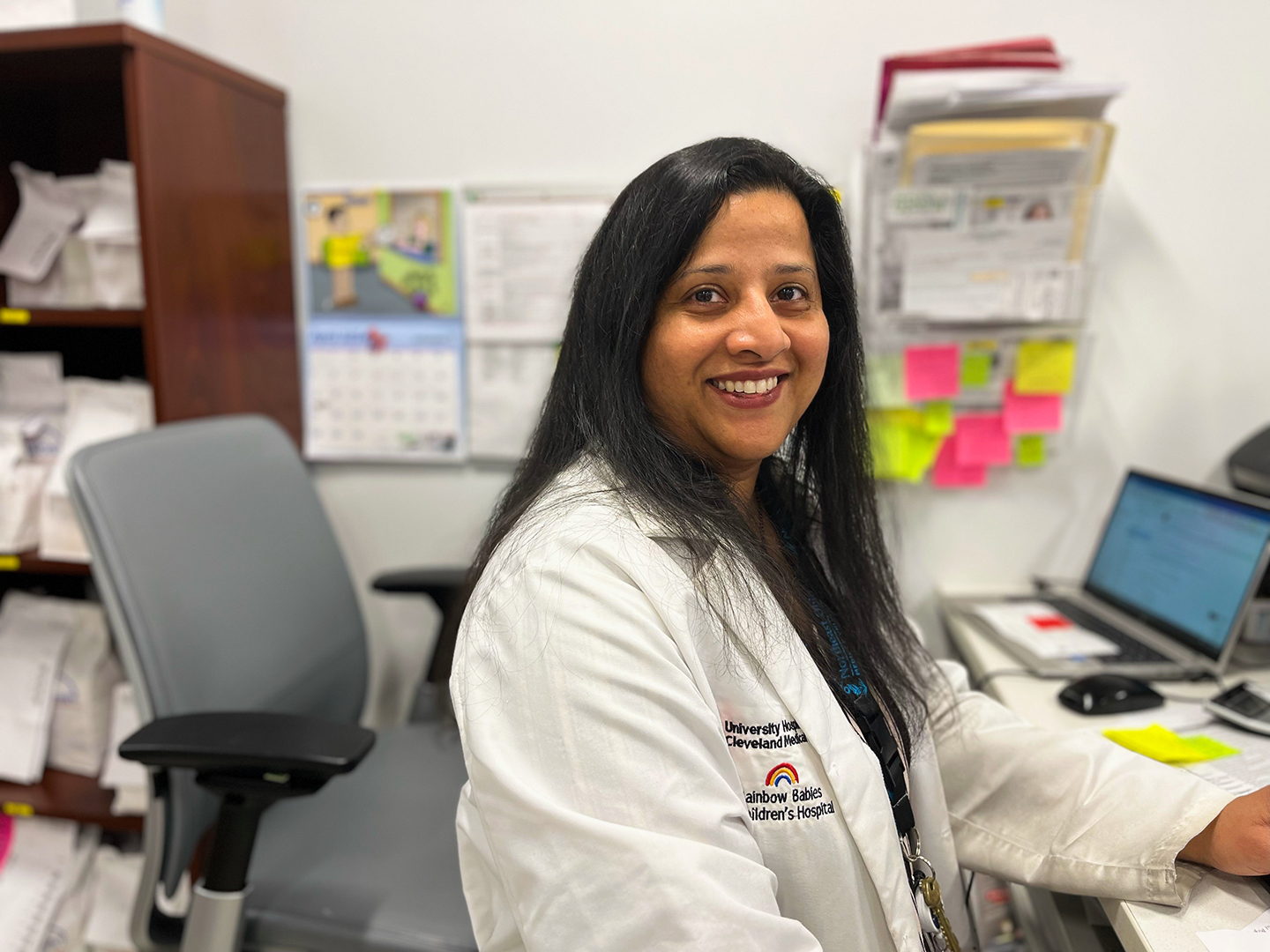 A photo of pharmacist Tanvi Jani sitting at her desk. She is wearing her white lab coat and has brown skin and long, dark hair.