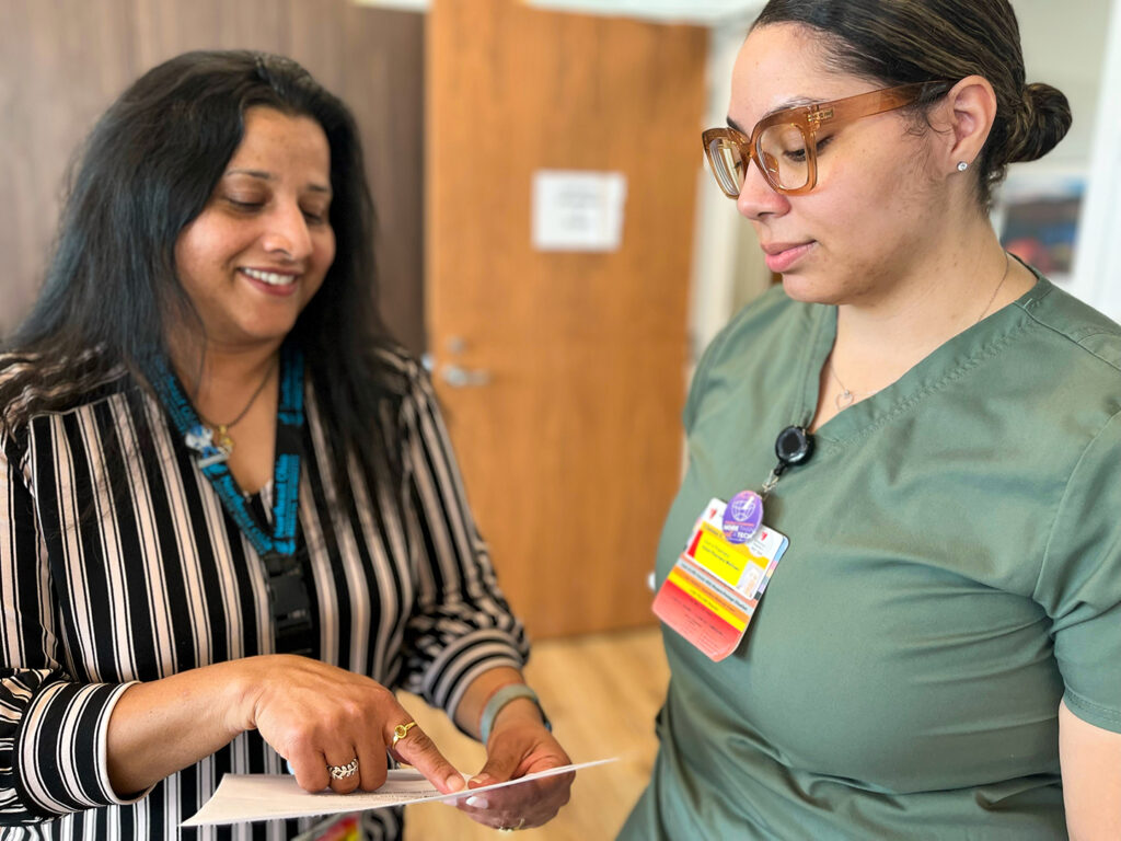 An image of pharmacist Tanvi Jani interacting with a nurse in green scrubs. She is discussing medication details while reading from a printed report.