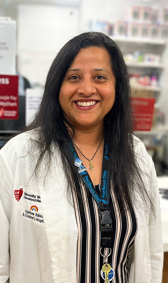 An image of pharmacist Tanvi Jani standing in front of a pharmacy counter. She is wearing a white lab coat over a black-and-white striped shirt.