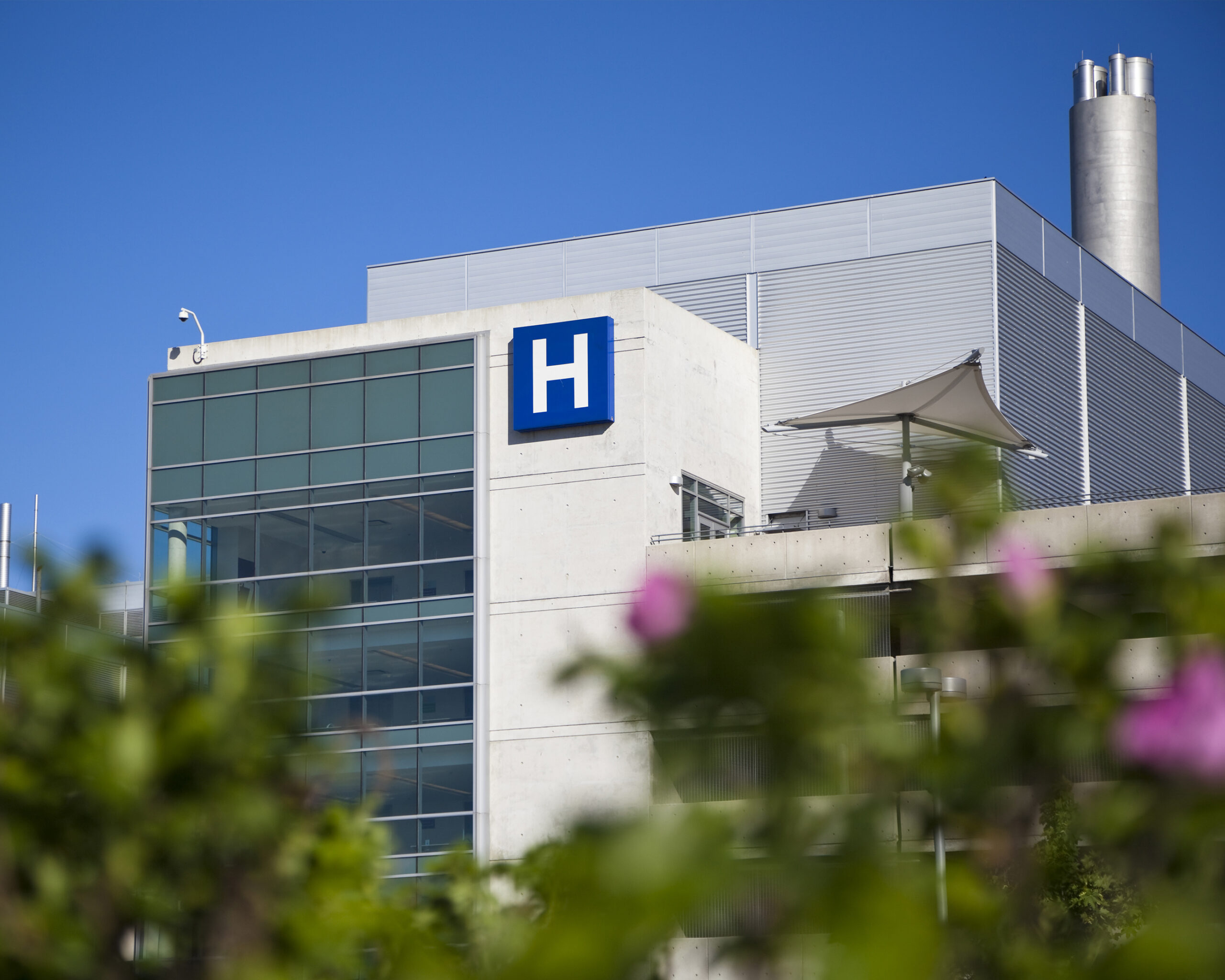 An exterior photo of a hospital. The building features walls of concrete and glass and a prominently placed H symbol.