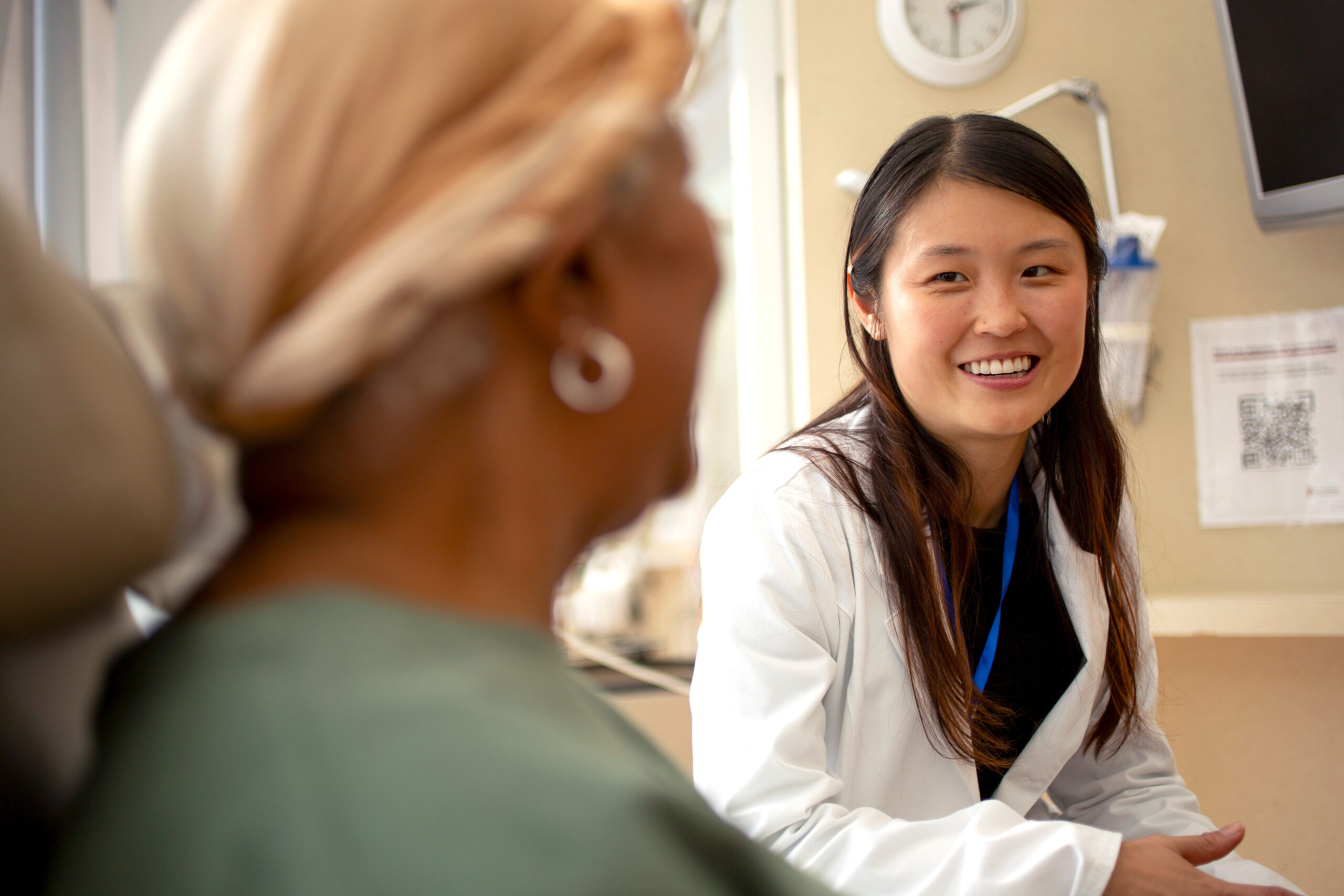A health-system pharmacist speaking to a patient. The pharmacist is an Asian woman with long, dark hair, and the patient is in the foreground, facing away from the camera.