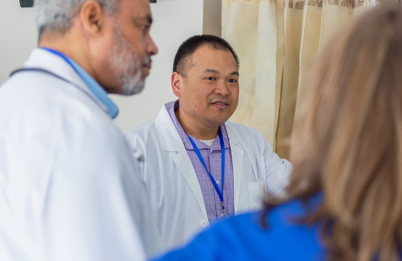 A health-system pharmacist speaking with other health care professionals. The pharmacist is an Asian man with short hair and wearing a white lab coat over a lavender dress shirt.