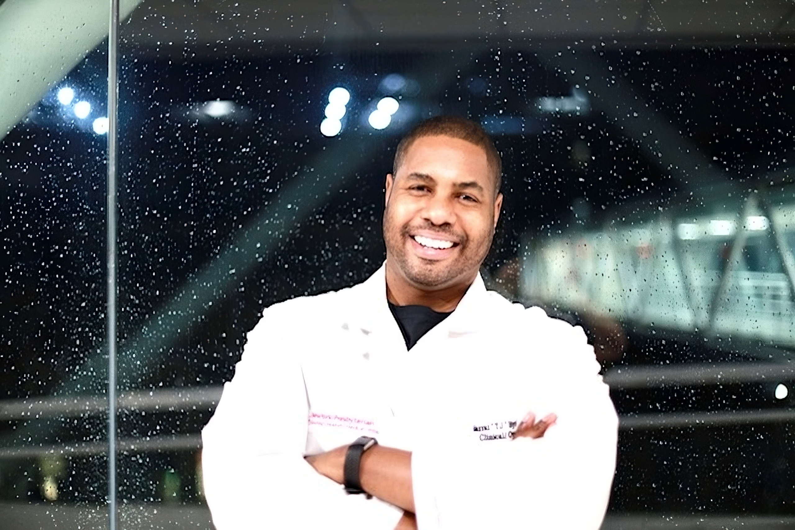 An image of pharmacist Charrai Byrd standing with his arms crossed in front of a rainy window at night. He is a Black man and is wearing a white lab coat.