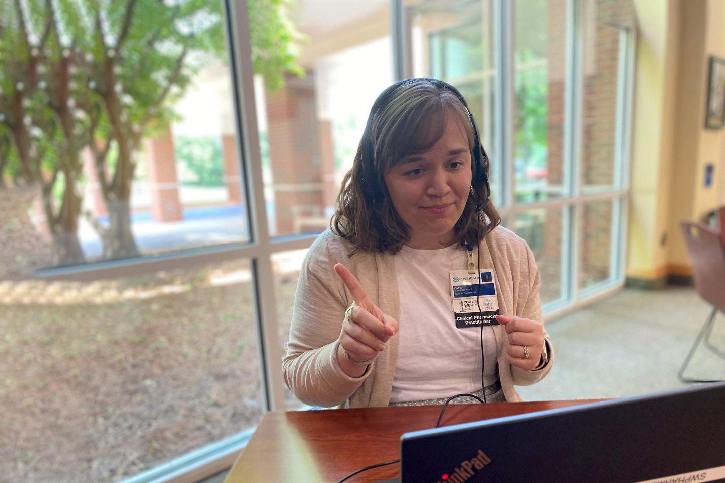 Pharmacist Catie Harper is talking on a video conference televisit. Harper is wearing a black headset with a microphone, a tan cardigan, and a hospital ID badge.