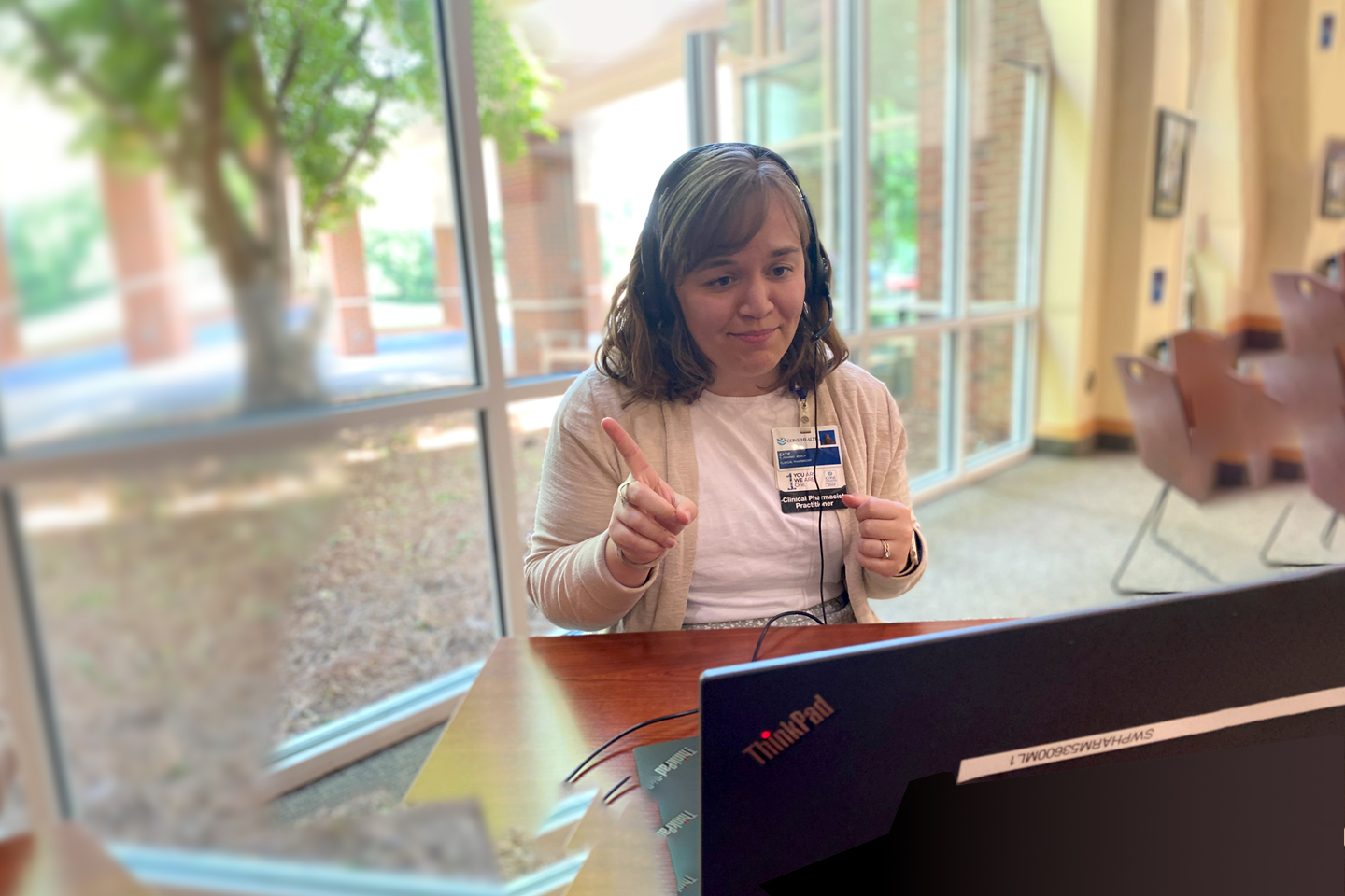 Pharmacist Catie Harper is talking on a video conference televisit. Harper is wearing a black headset with a microphone, a tan cardigan, and a hospital ID badge.