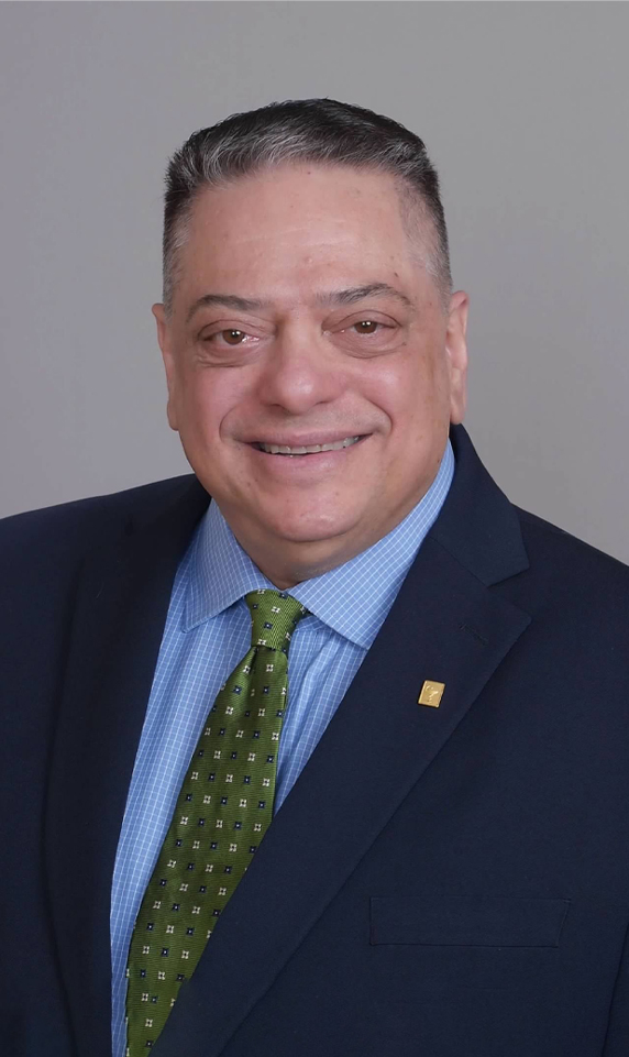 A headshot-style image of pharmacist Gene B. Decaminada. He is wearing a dark blue suit, light blue shirt and green tie.