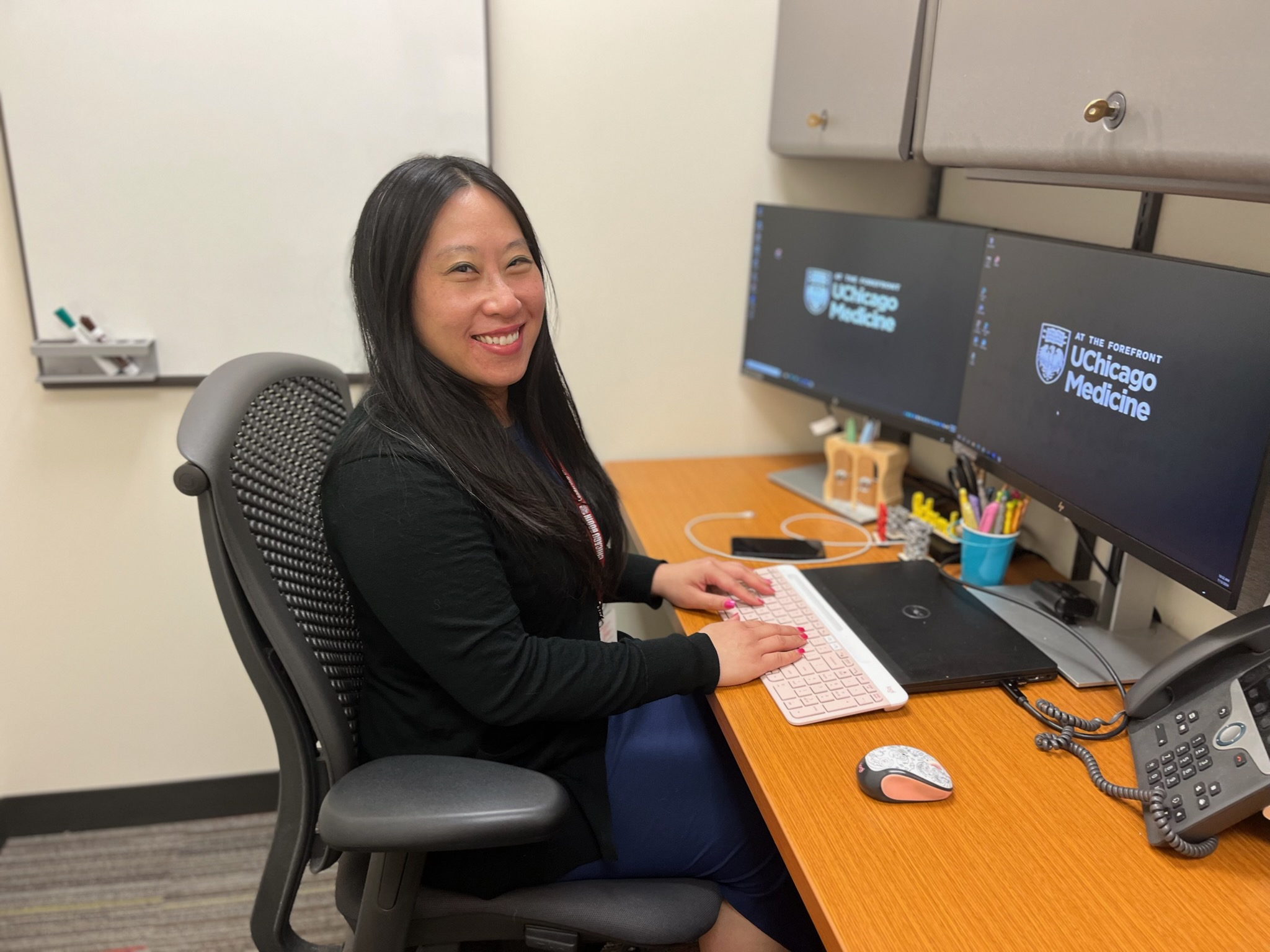 Pharmacist Bernice Man is smiling - she is sitting at her desk in front of her computer and two monitors.