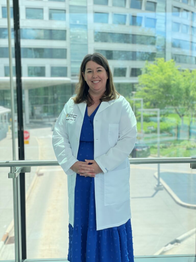 Pharmacist Kelly Hoenig is smiling - she has a white lab coat on and is standing in front of a window.