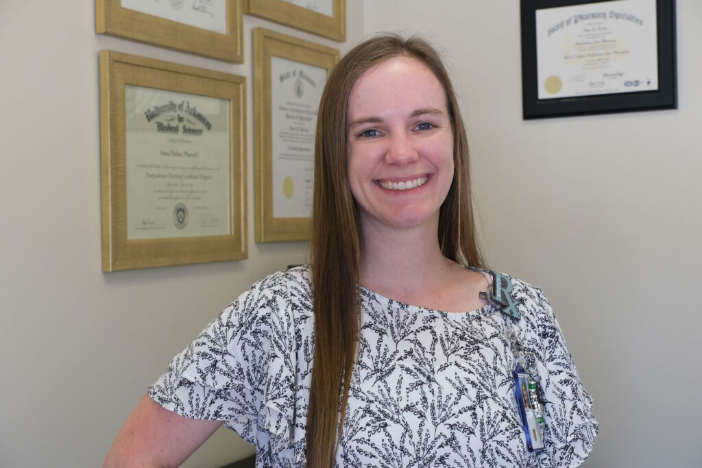 Anna Parker stands in her office, with her medical diplomas and board certifications hanging on the walls behind her. She is smiling at the camera and wearing her work RX badge and ID.