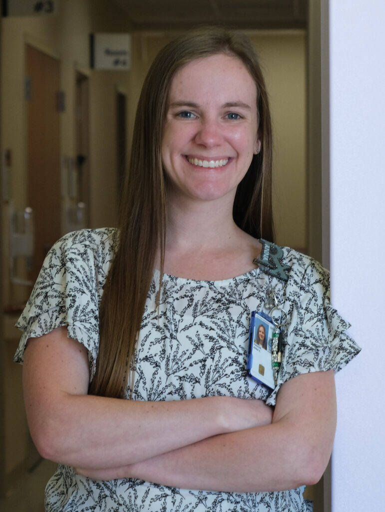 Anna Parker smiles at the camera in the hallway of a hospital. She is wearing her RX ID badge and is ready to help patients with medication questions.