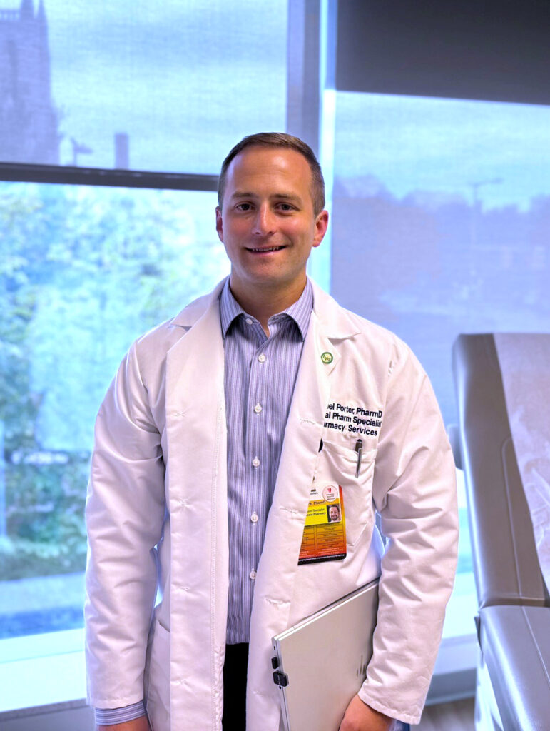 Samuel Porter, standing in an exam room in a lab coat with laptop in hand. Windows and exam table are in the background.