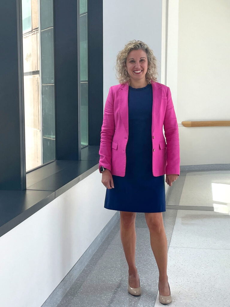 Tanya is standing in the hall of Penn Medicine next to a row of windows in a pink suit jacket and blue dress.