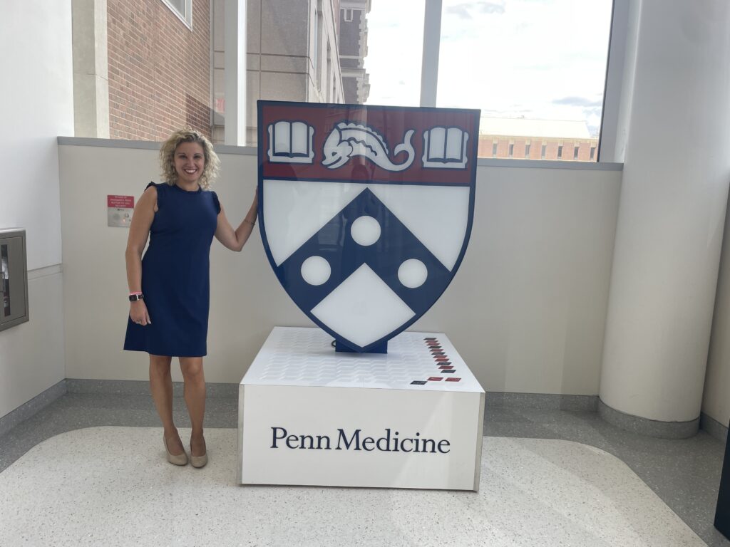 Tanya is standing next to Penn Medicine Logo in the medical school hallway.